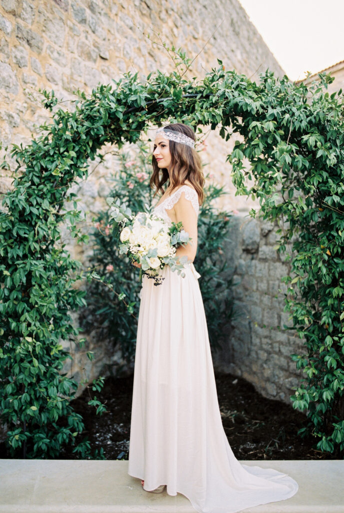Bride with a bouquet stands near an arch of green ivy against a stone wall. High quality photo