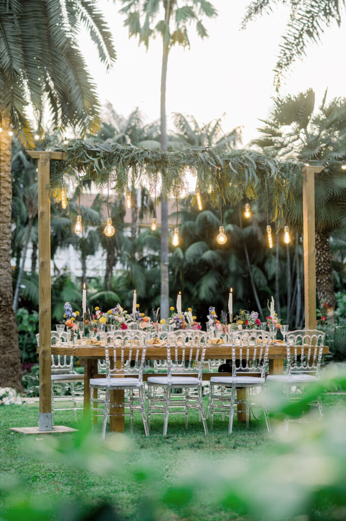 Wooden table with arch decorated with plants and hanging light bulbs and transparent chairs ready for wedding banquet in tropical resort